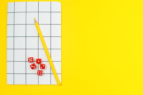 Yellow Colored Pencil and Red Dice on Notebook