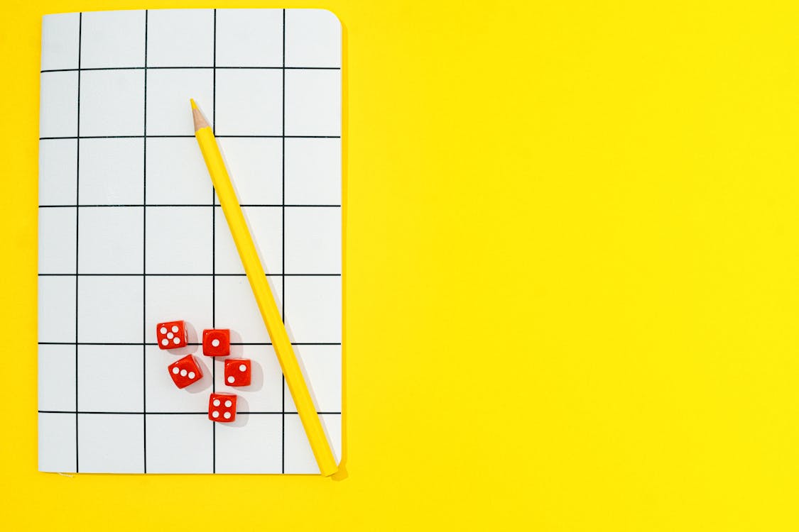 Yellow Colored Pencil and Red Dice on Notebook