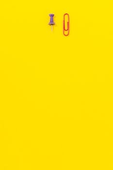 A minimalist bright yellow background featuring a red paper clip and purple push pin. Perfect for creative designs.