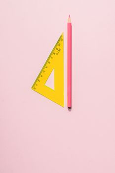 A vertical shot of a pink pencil and yellow triangular ruler on a pastel pink surface.