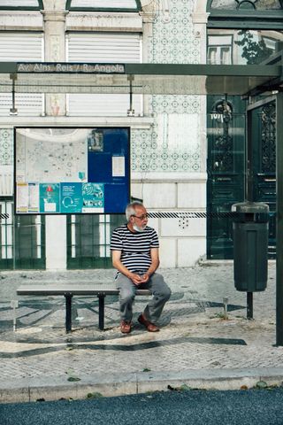 Senior man sitting at a Lisbon bus stop, wearing a mask and striped shirt, capturing urban lifestyle.