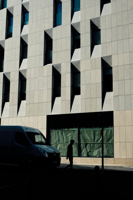 Shadowy figure walking near a geometric facade in Lisbon, under a van's silhouette.