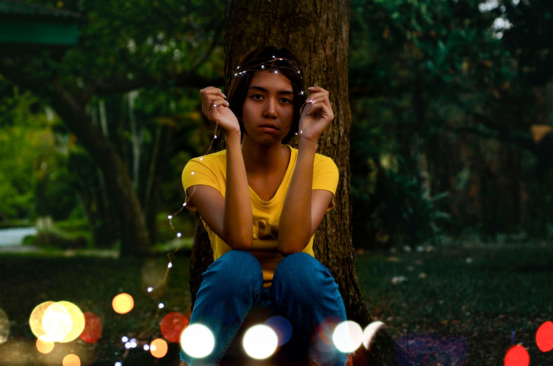 A young woman sits by a tree with twinkling fairy lights at dusk, embracing outdoor tranquility.