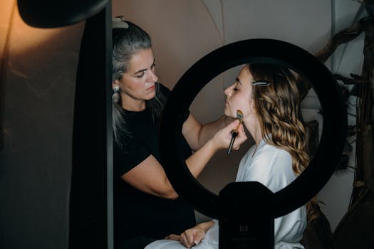 A makeup artist skillfully applies cosmetics to a woman under a ring light in an indoor setting.