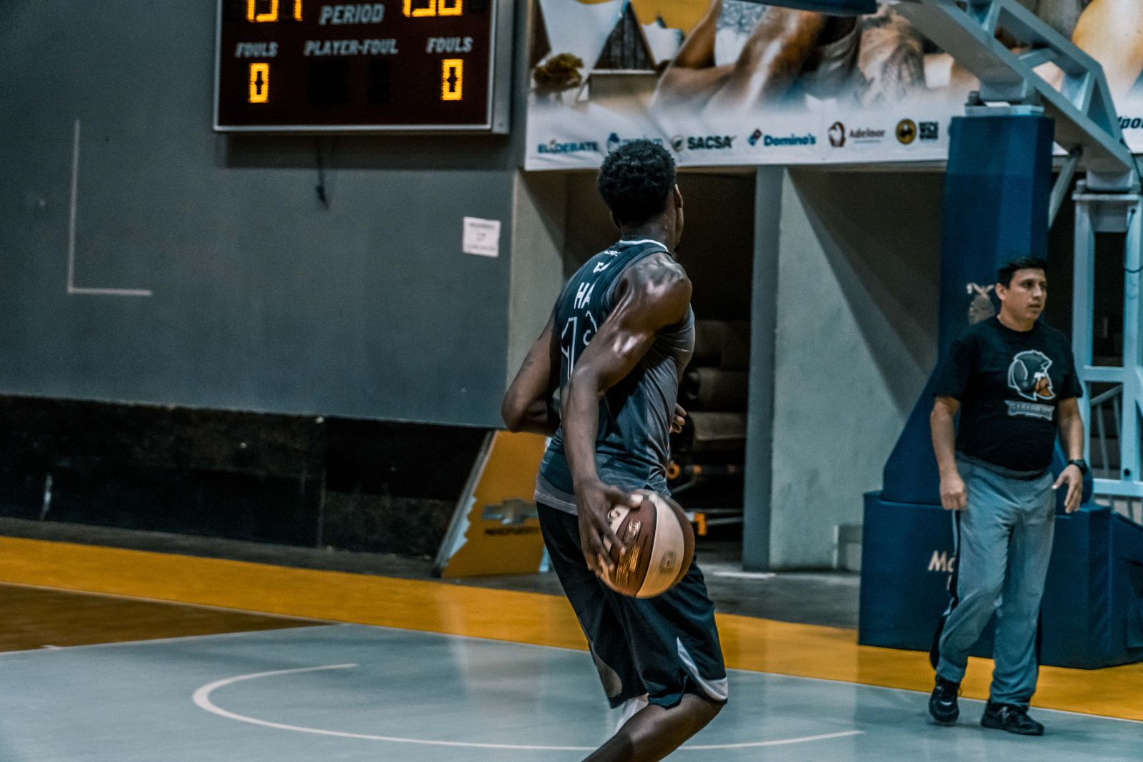 Jogador com a bola em quadra coberta, com o placar eletrônico visível ao fundo