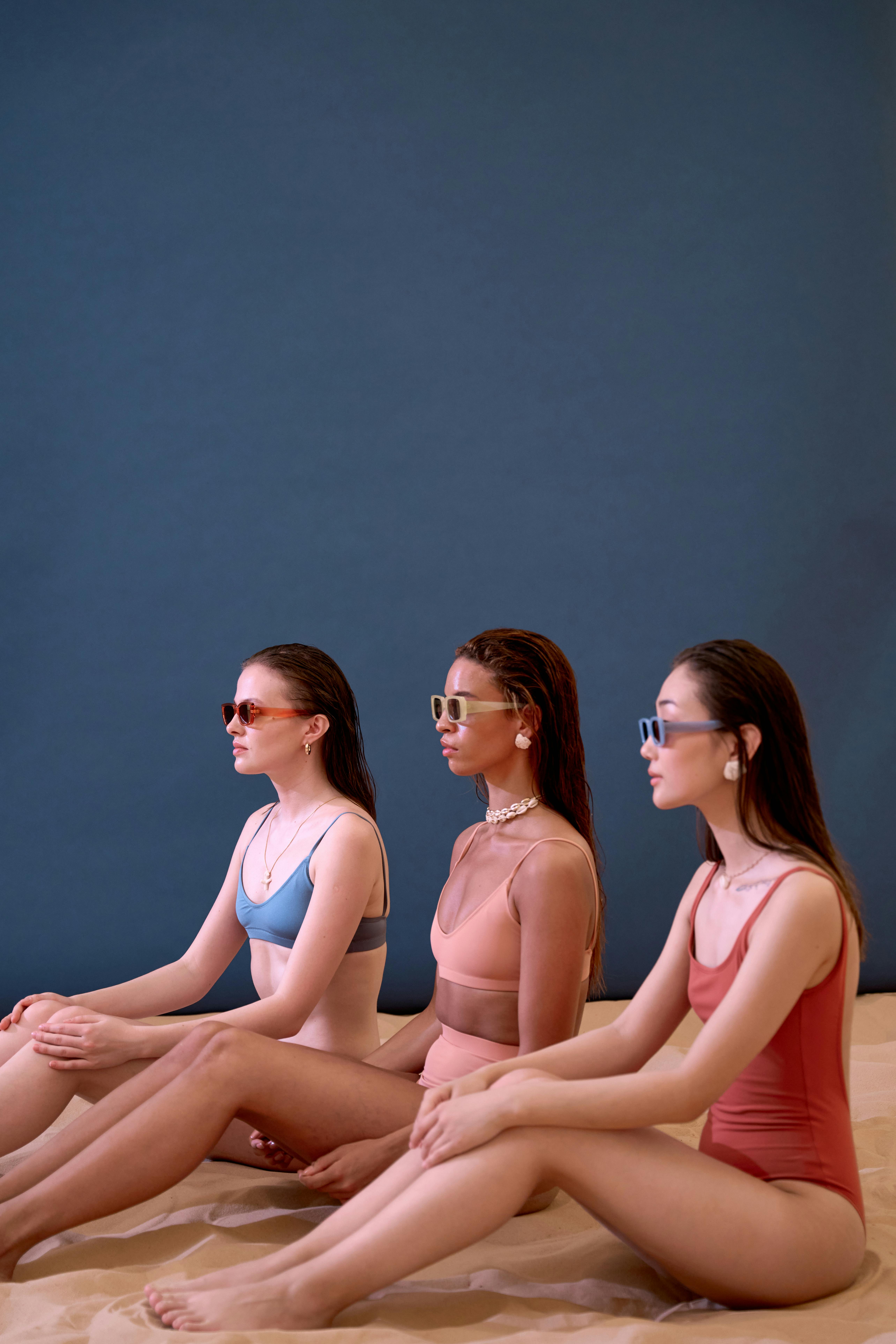 Three women wearing swimsuits and sunglasses sitting on brown sand with a blue backdrop.