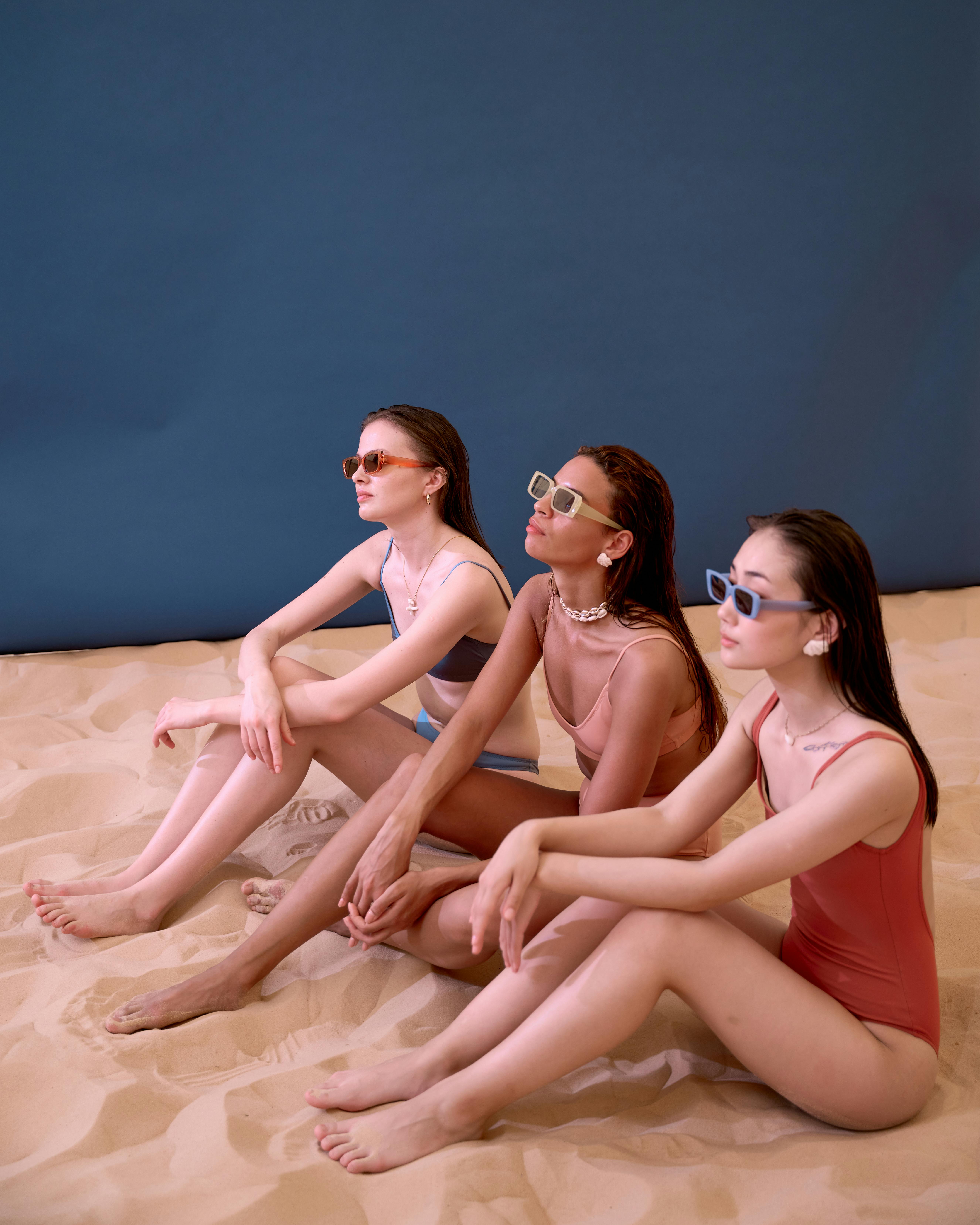 Three diverse women in stylish swimwear and sunglasses posing elegantly on sandy backdrop, showcasing fashion and diversity.