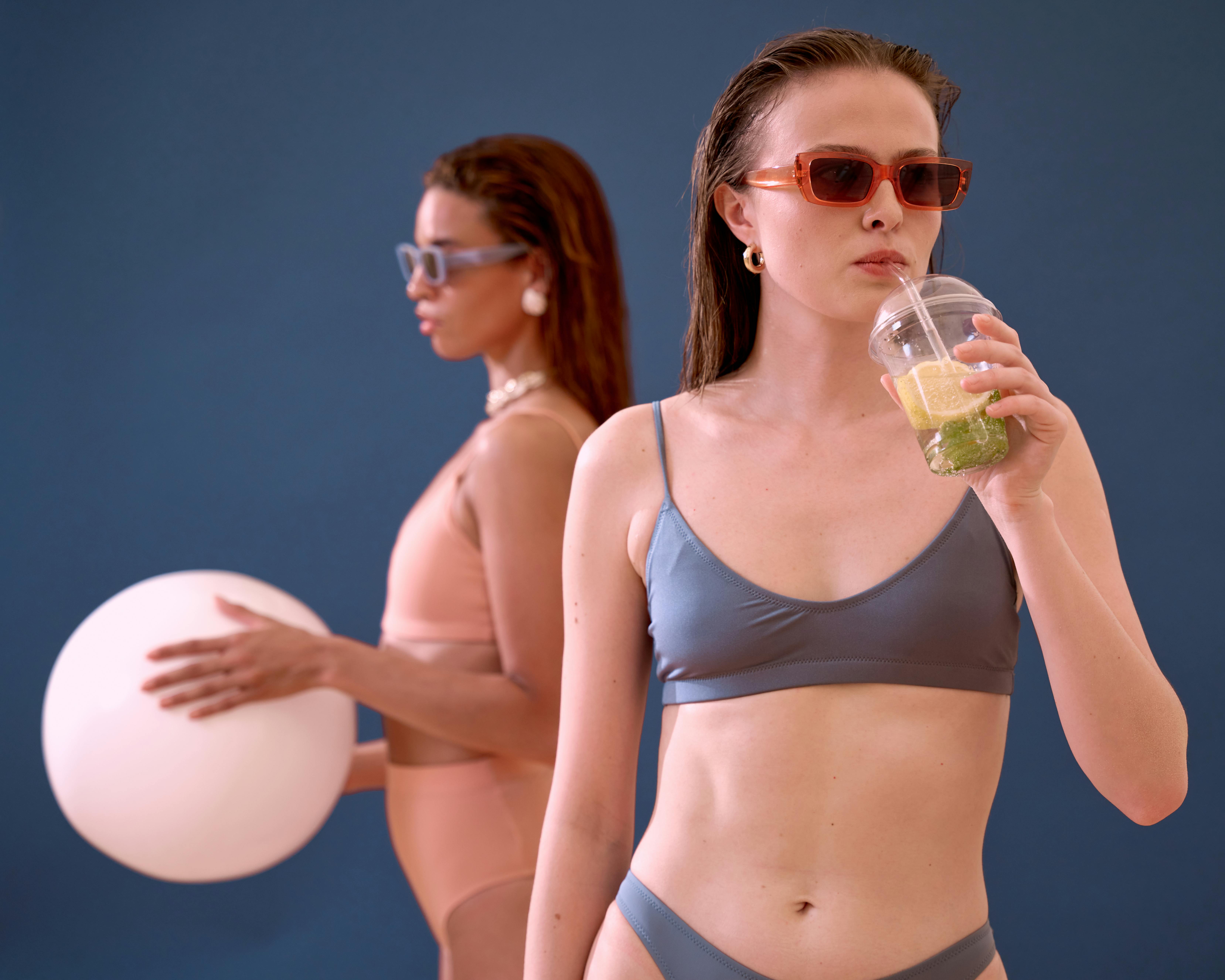 Two women in stylish bikinis enjoying summer vibes with drinks and beach accessories.