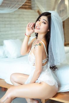 Asian woman in bridal lingerie with a veil, tattooed and smiling indoors.
