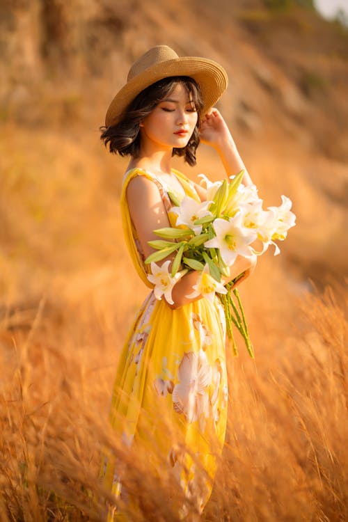 Shallow Focus of a Beautiful Woman in Yellow Floral Dress Standing on Grass Field while Holding Flowers