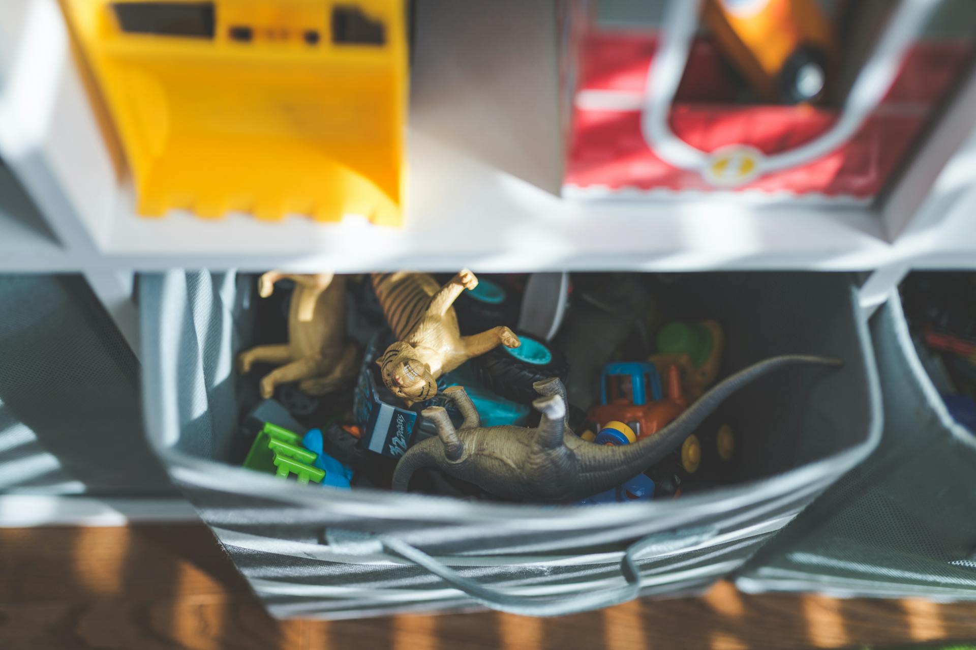 Plastic dinosaurs and cars in a storage box in a sunlit room.