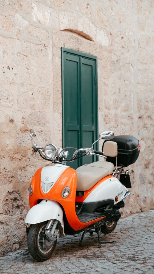 Free A retro orange and white scooter parked in a quaint alley against a stone wall. Stock Photo