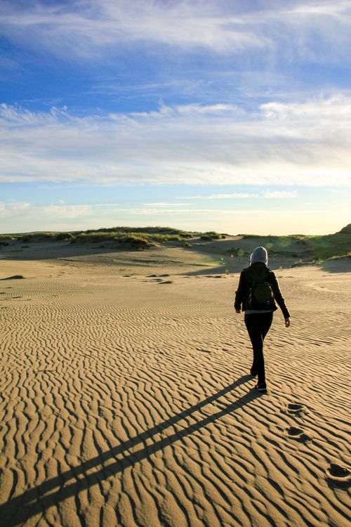 Free A solitary figure walks across sunlit sand dunes under a vibrant blue sky. Stock Photo