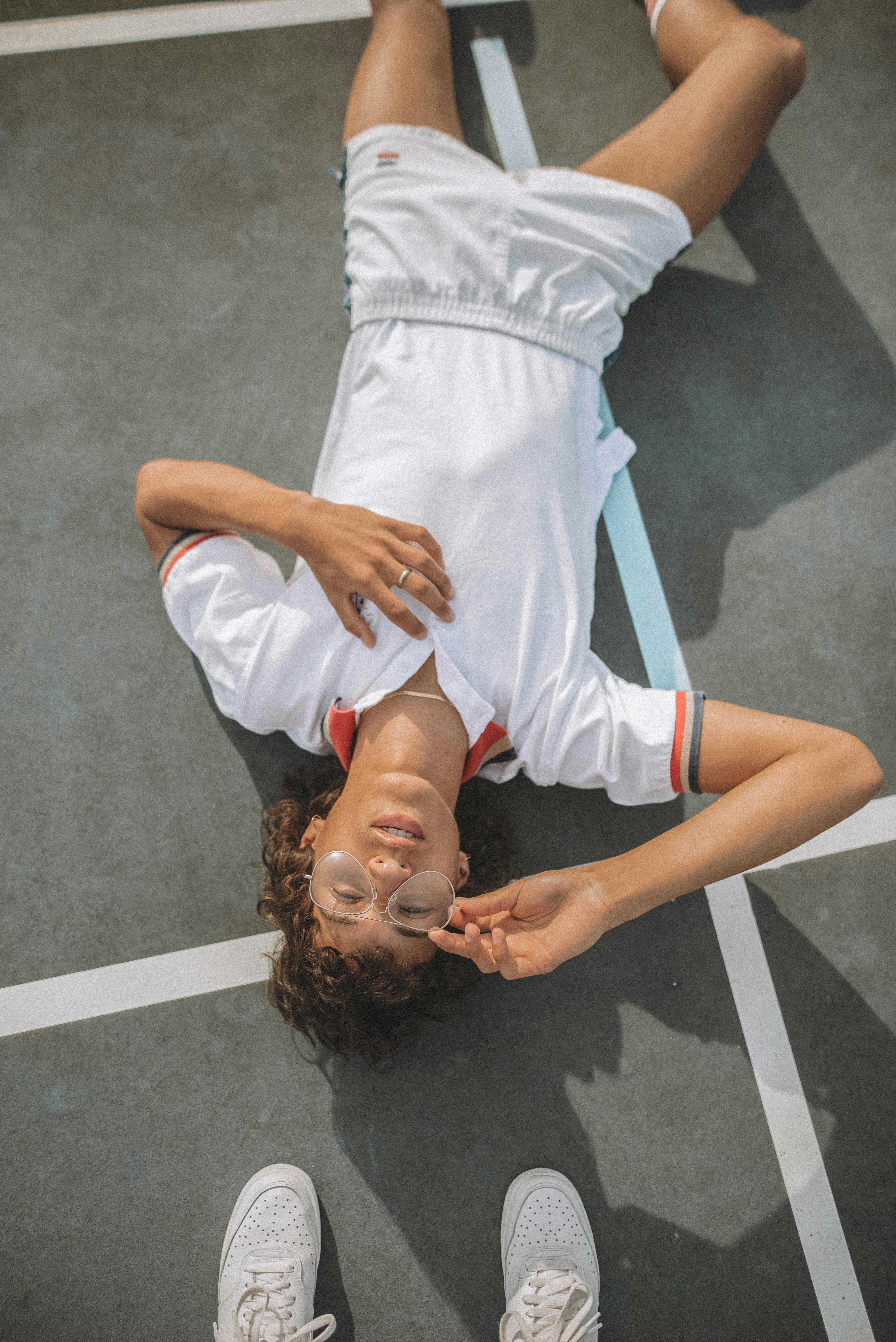 A young adult with curly hair lies on a sports court in casual attire, wearing eyeglasses.