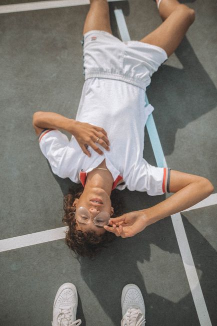 A young adult with curly hair lies on a sports court in casual attire, wearing eyeglasses.