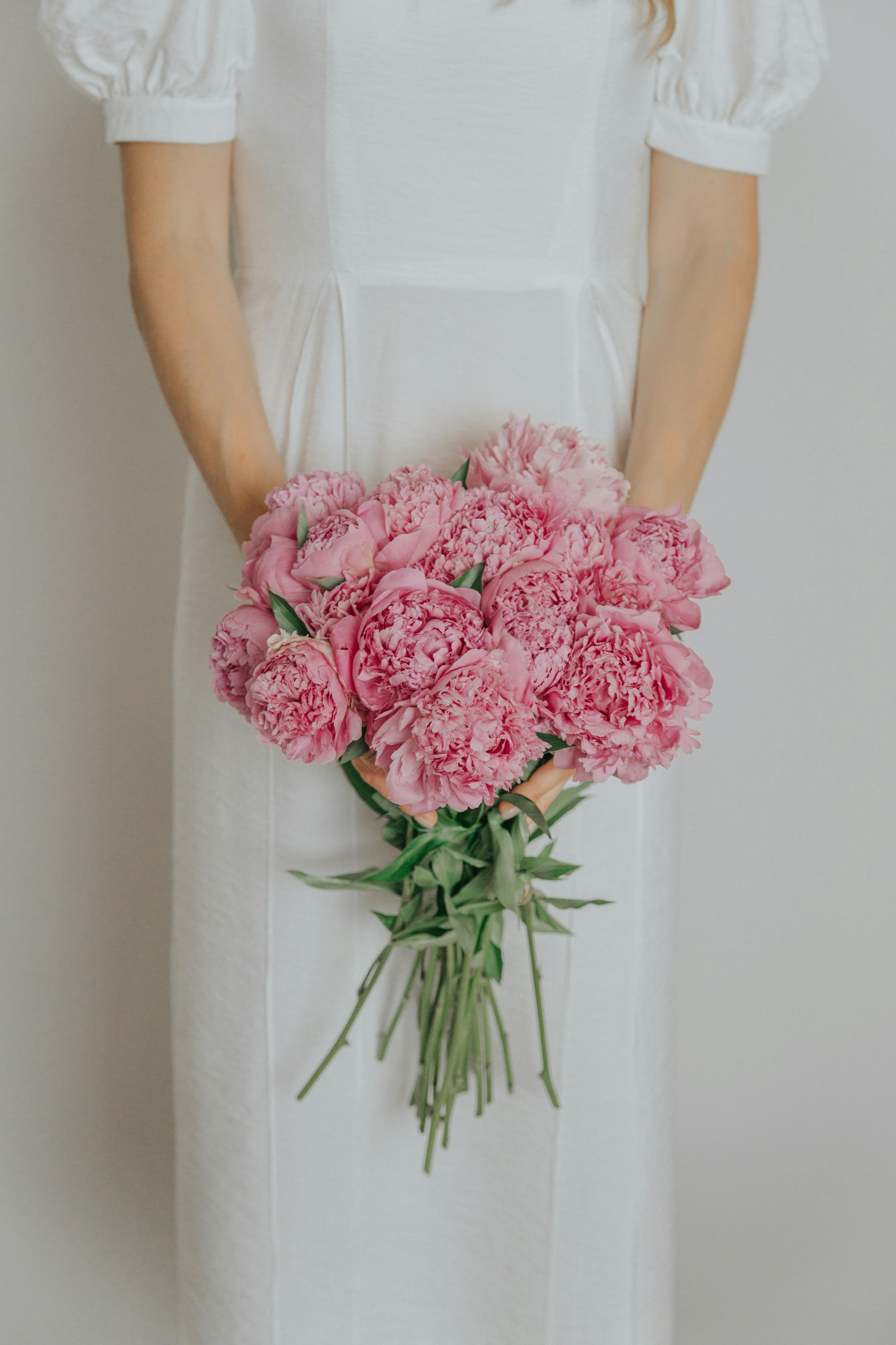 A woman in a white dress gently holding a bouquet of fresh pink peonies against a white background.