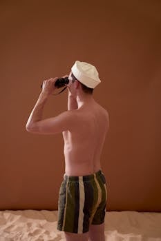 Back view of shirtless man wearing a sailor hat and shorts using binoculars indoors.