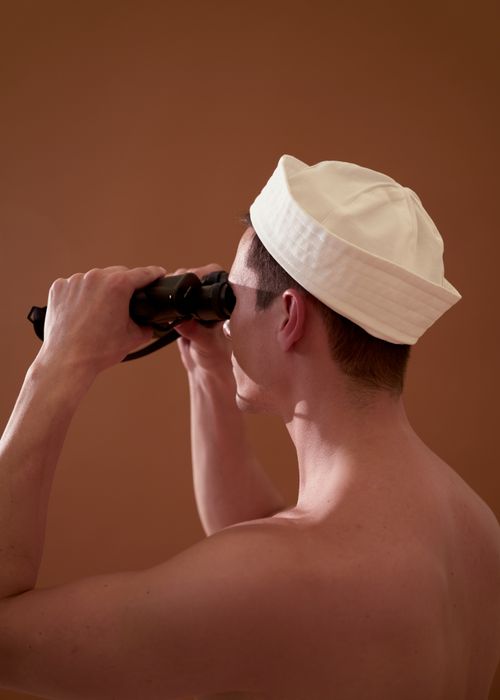 Free A shirtless man in a white hat uses binoculars against a brown background. Stock Photo