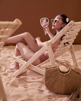 Asian woman relaxing in swimwear on a beach chair with a handheld mirror and straw bag.