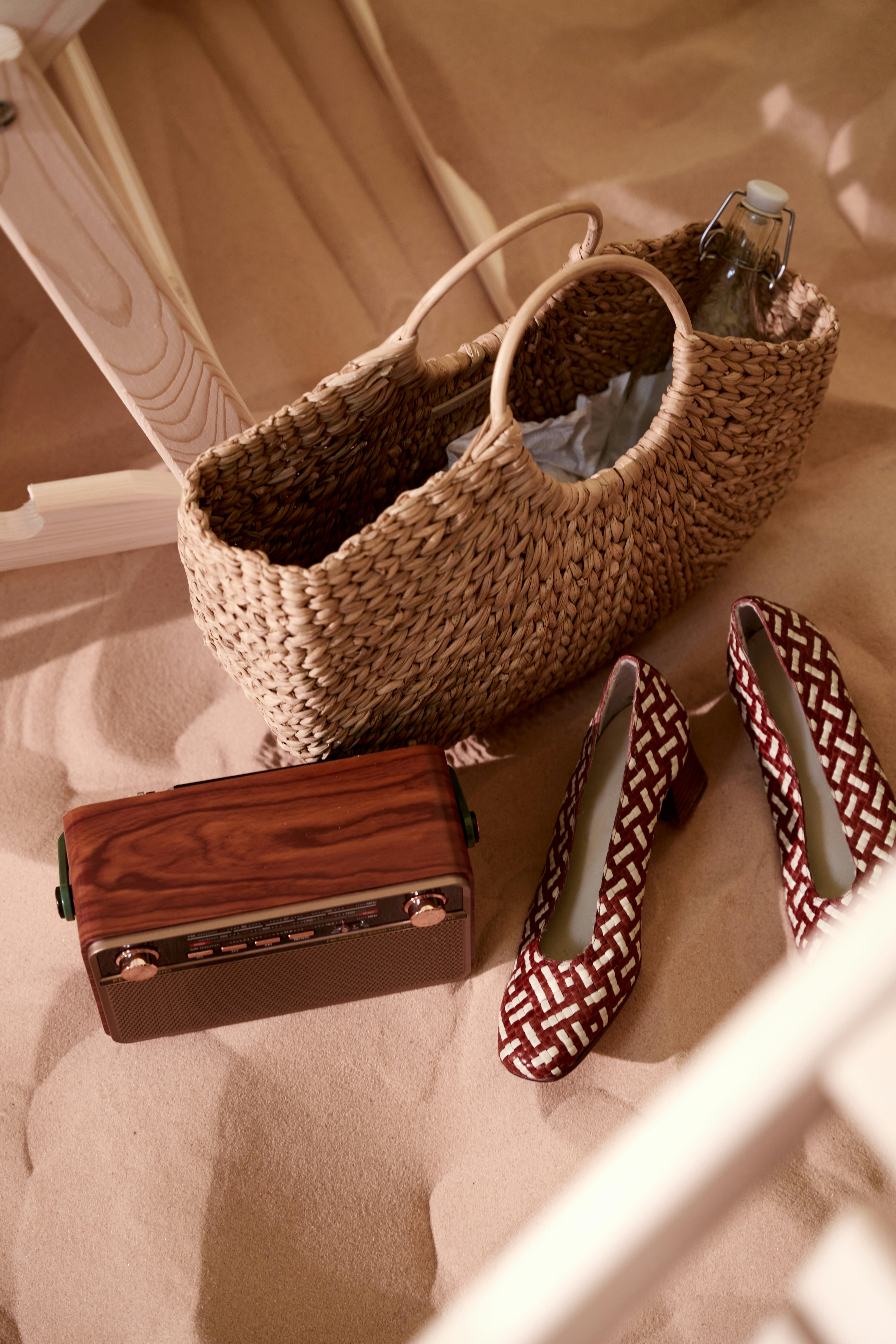 A stylish arrangement featuring a straw bag, retro radio, and patterned shoes on sand.