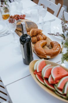 An elegant outdoor dining table set with wine, bread, croissants, and watermelon slices, perfect for a celebration.