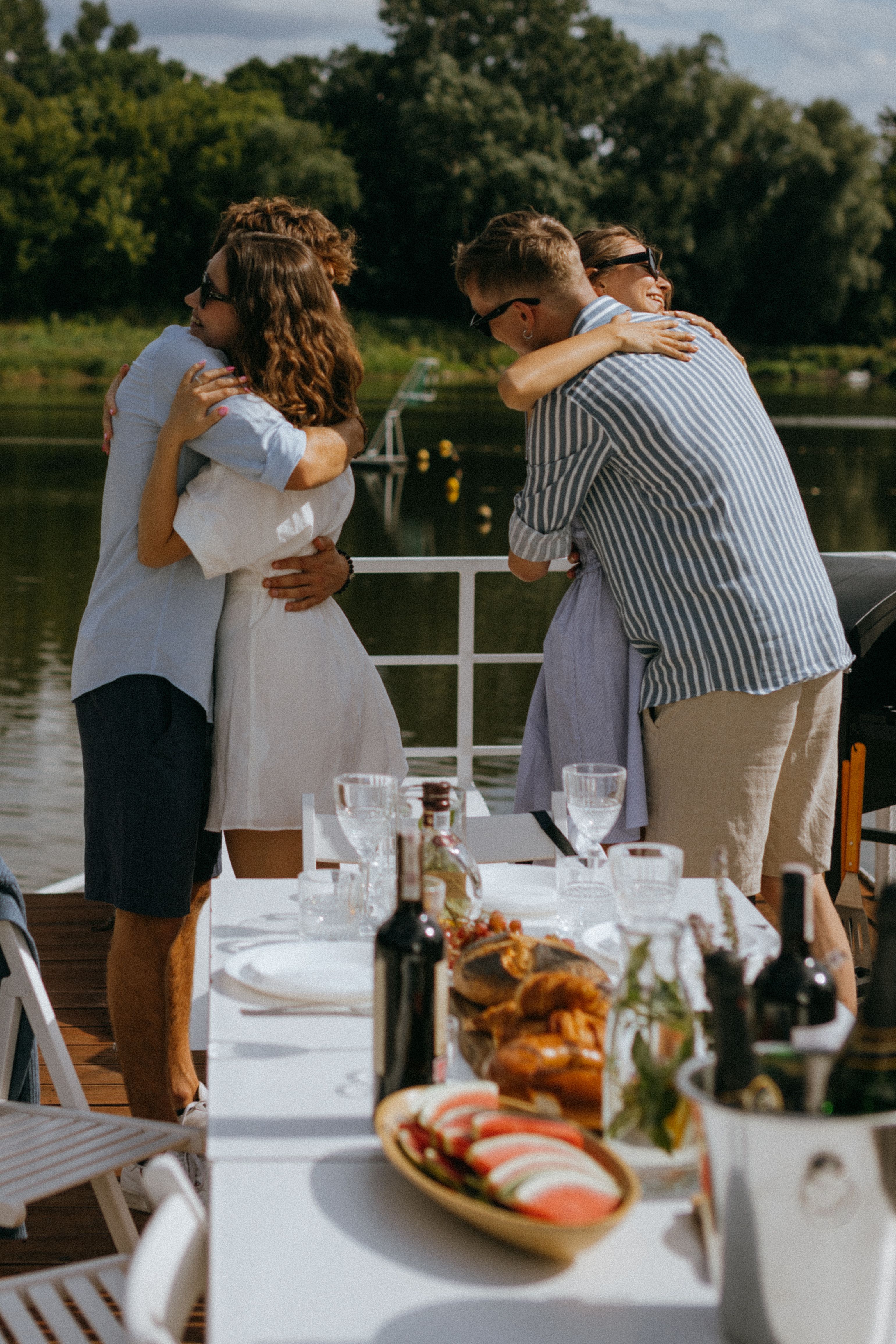 A joyful outdoor gathering by a lake with friends hugging and a table set for a meal.