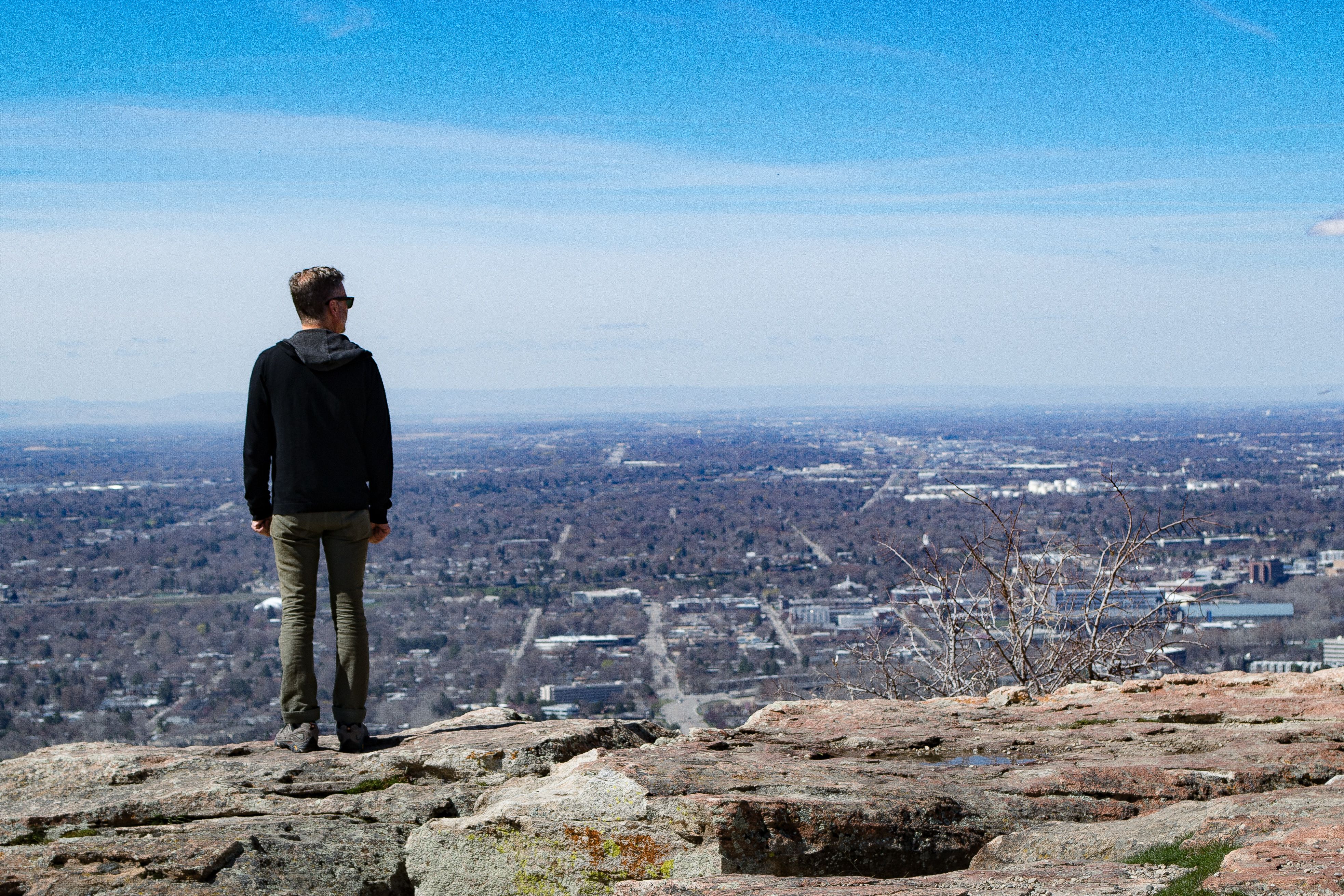 A man stands on a cliff edge, taking in a vast urban landscape below on a sunny day.