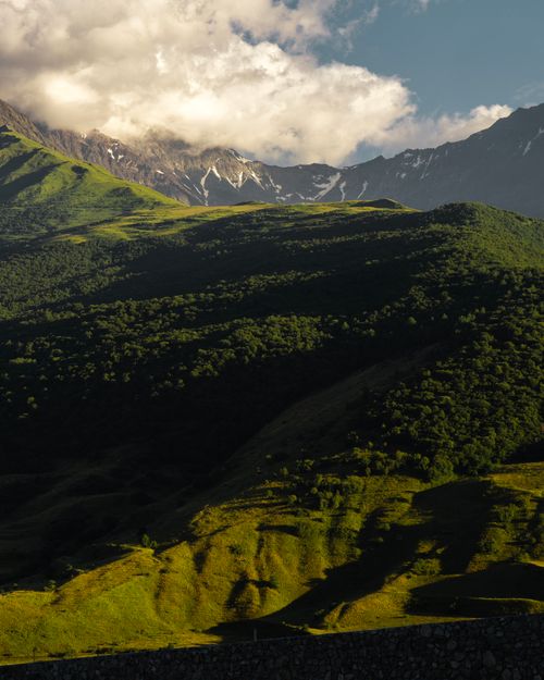 Free Scenic view of sunlit mountains and lush forests under a dramatic cloudy sky. Stock Photo
