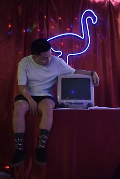 A young man sits by a vintage television under a dinosaur neon light.