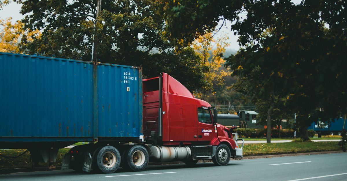 A red truck transports a blue cargo container on a scenic, tree-lined road.
