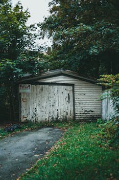 Old wooden garage with 'For Sale' sign surrounded by overgrown greenery.