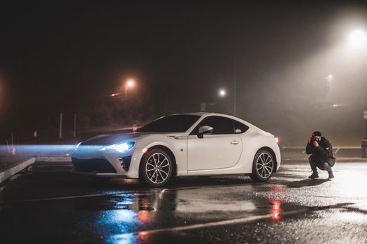 A white coupe in a dimly lit parking lot with a photographer capturing the moment at night.