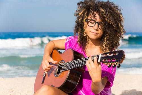 Woman Wearing Purple Shirt Playing Brown Classical Guitar While Sitting Near Shoreline With Water Splashing Background in Daytime