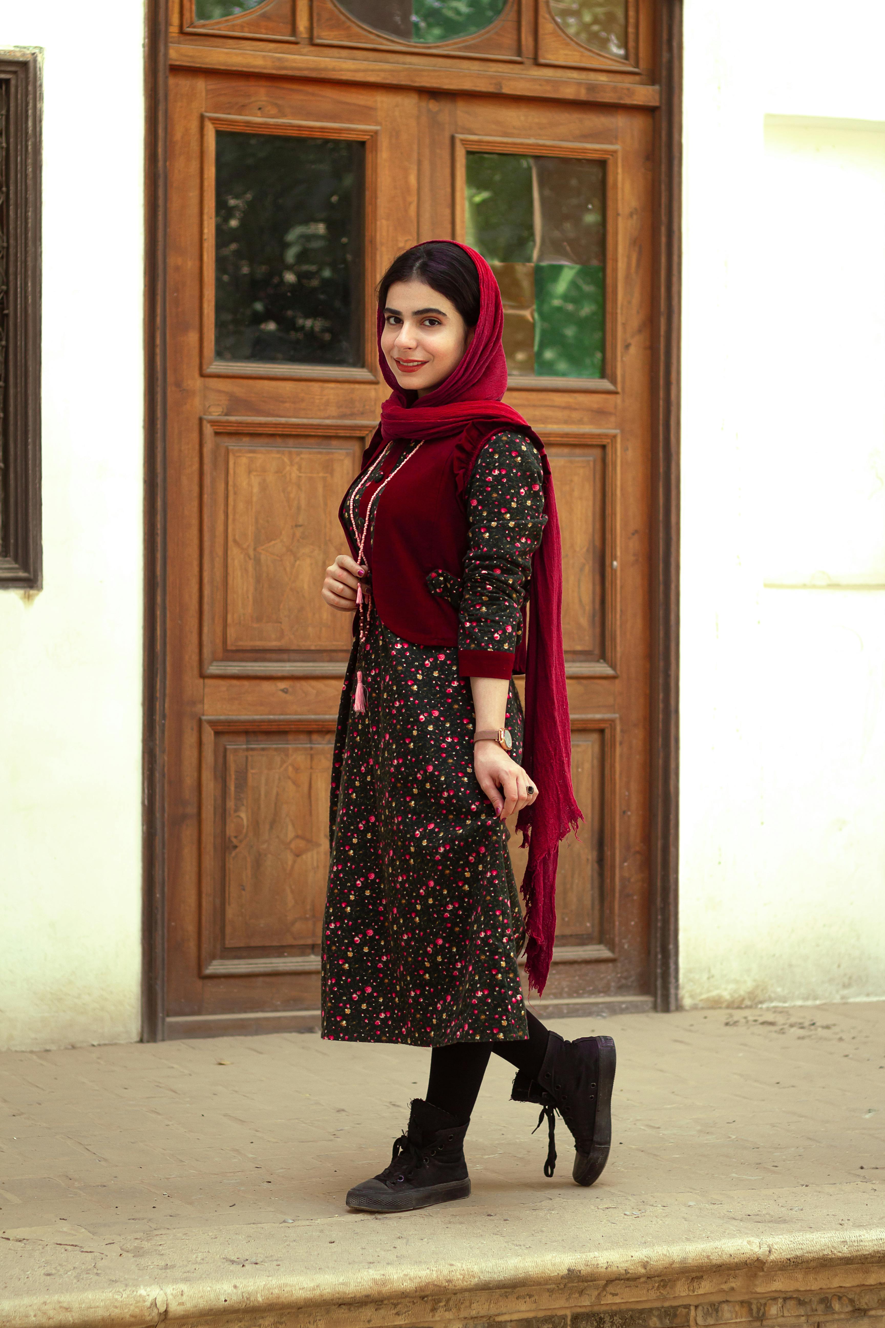 Woman in elegant floral dress and hijab standing by a wooden door.