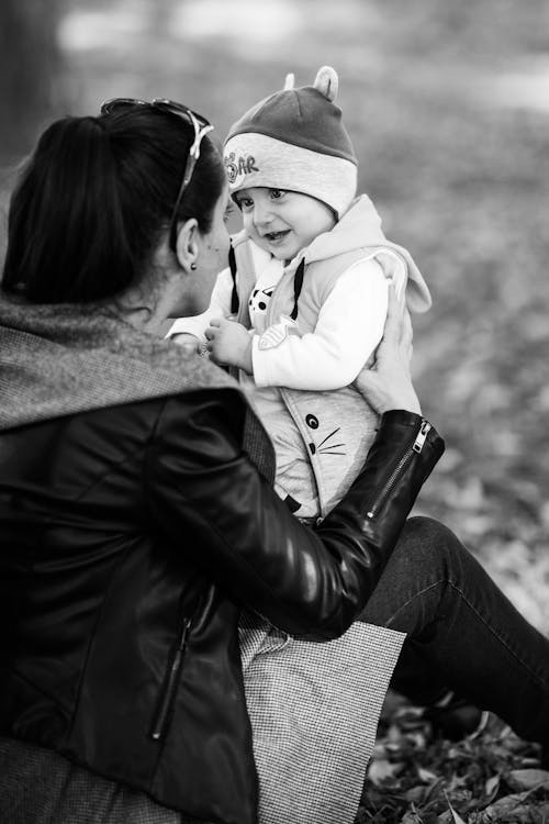 Grayscale Photo of Woman in Leather Jacket Holding a Baby on her Lap