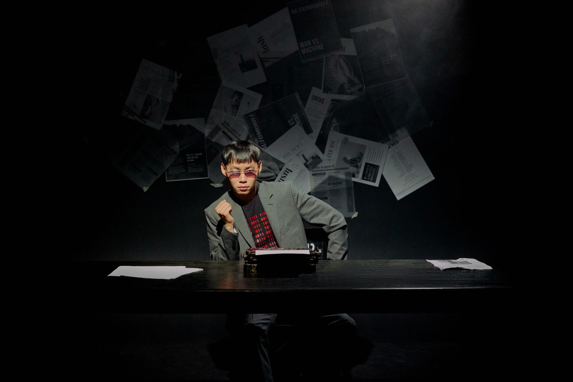 Man in sunglasses typing on a typewriter in a dimly lit room surrounded by papers.