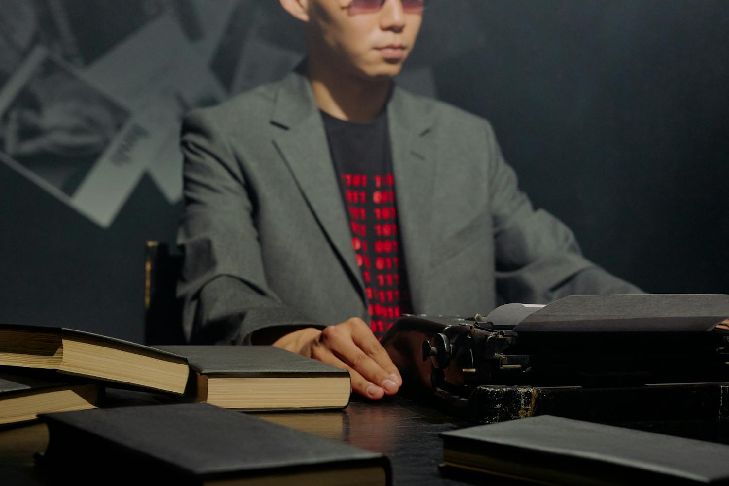 An Asian man in a gray blazer typing on a classic typewriter surrounded by books.
