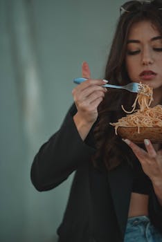 A young woman eats spaghetti from a glass bowl with a fork, capturing a casual dining moment.