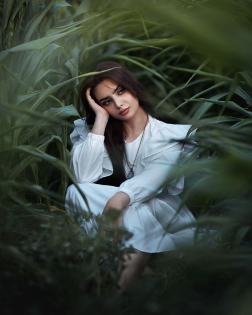 Free Elegant woman in white dress sitting thoughtfully amidst lush greenery, capturing a serene and introspective mood. Stock Photo