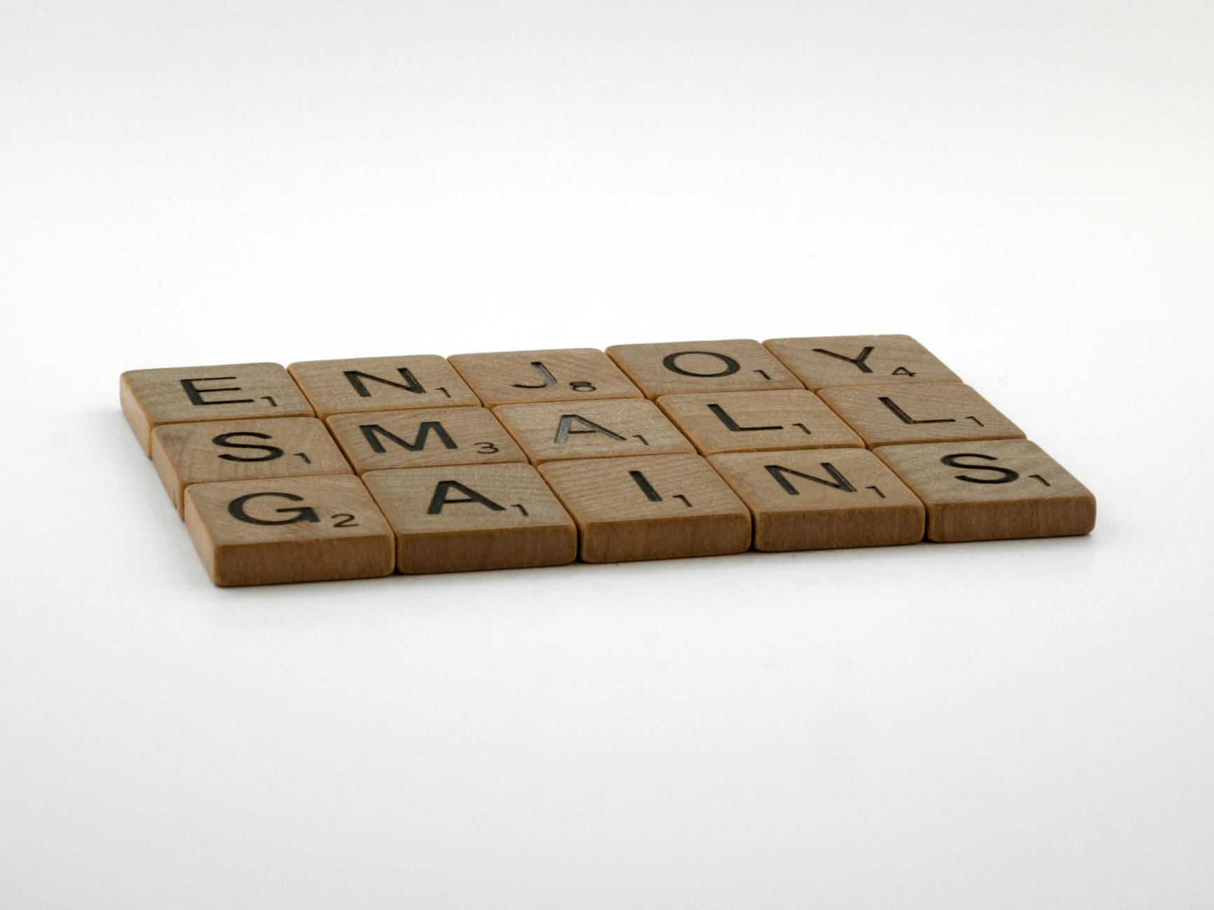 Close-up of Scrabble tiles forming the phrase 'Enjoy Small Gains' on a neutral background.
