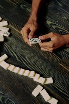 A close-up photo of hands holding white dominoes on a dark wooden table.