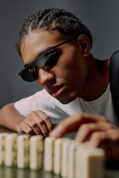 A fashionable man wearing sunglasses setting up domino blocks on a studio table.