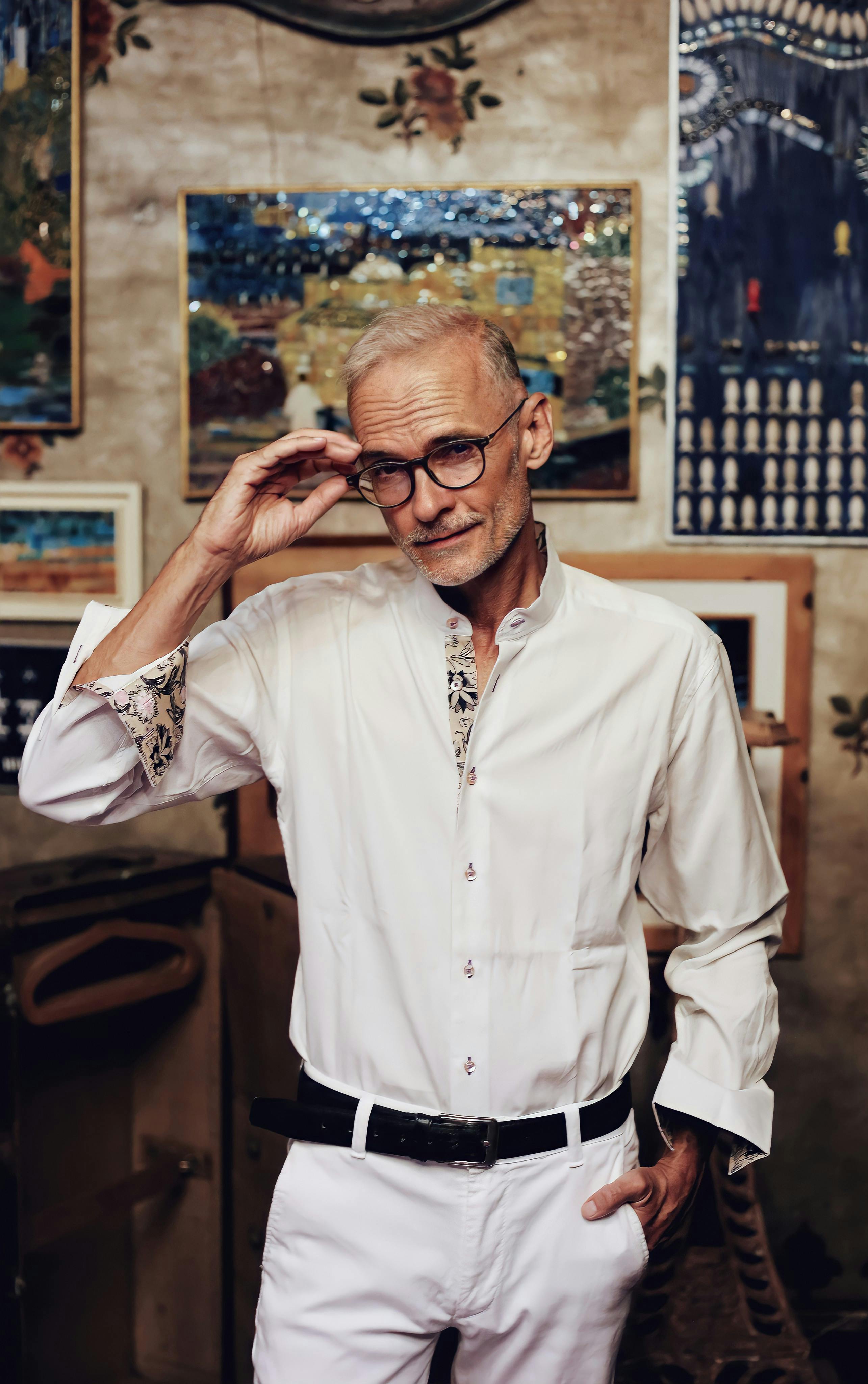 Senior man in white attire posing with glasses in an art-filled room.