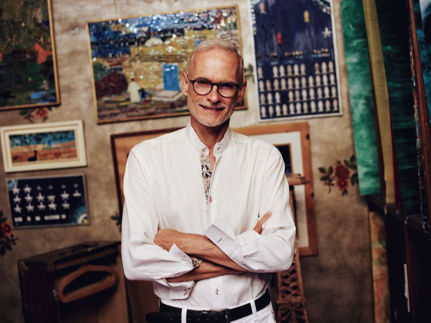 A senior man in glasses smiling while posing in an art gallery filled with paintings.