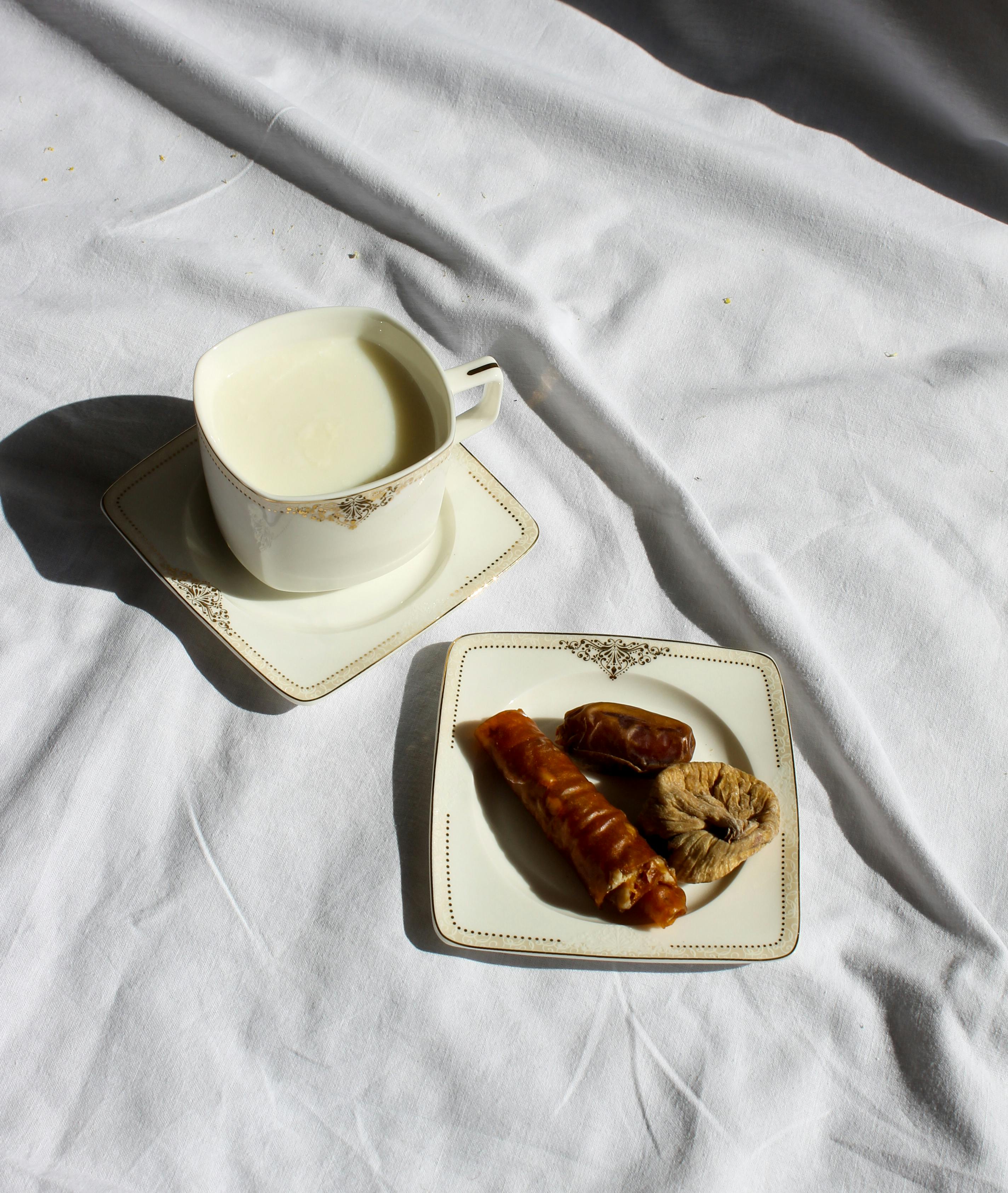 A cup of coffee on a saucer next to a plate of dried fruits atop a white linen tablecloth.