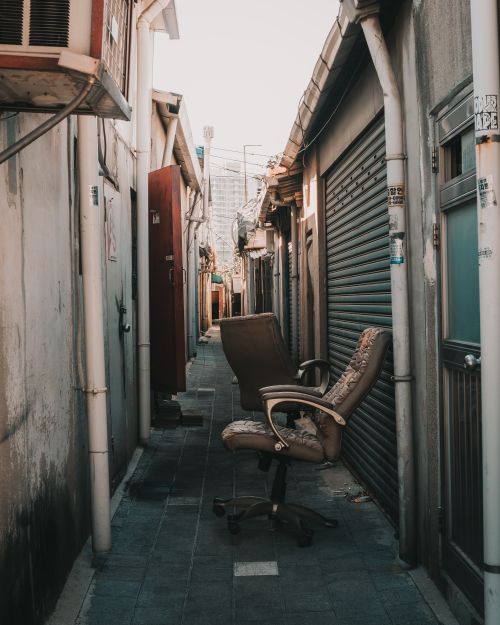 Free A narrow urban alleyway featuring office chairs and a roll-up door in daylight. Stock Photo