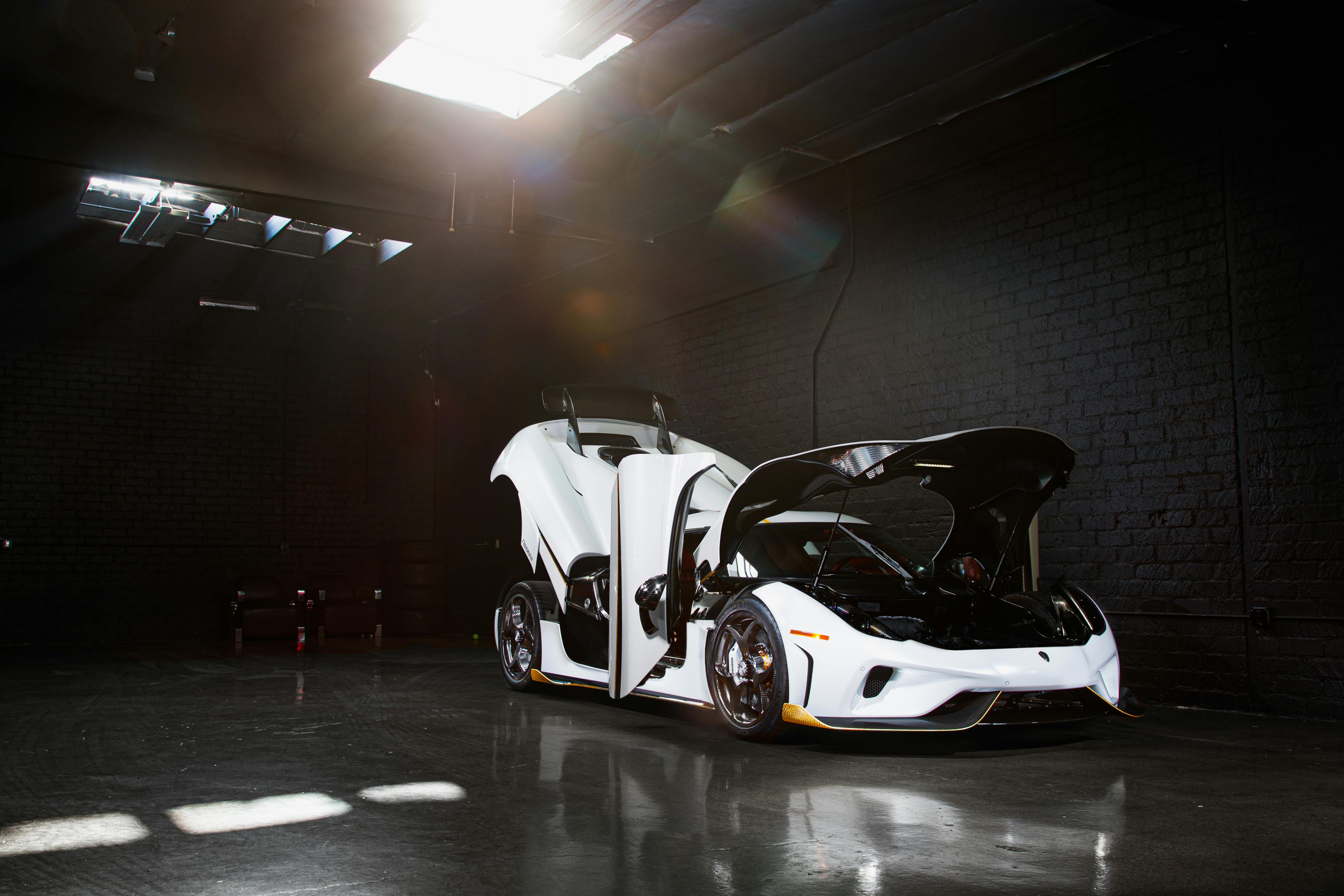 A white luxury sports car with gullwing doors open, showcased in a dark garage setting.