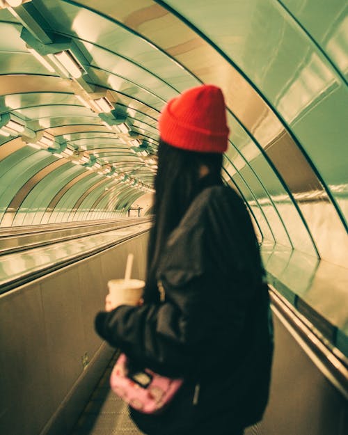Free A Woman in Black Jacket and Red Bonnet Standing on the Escalator Stock Photo