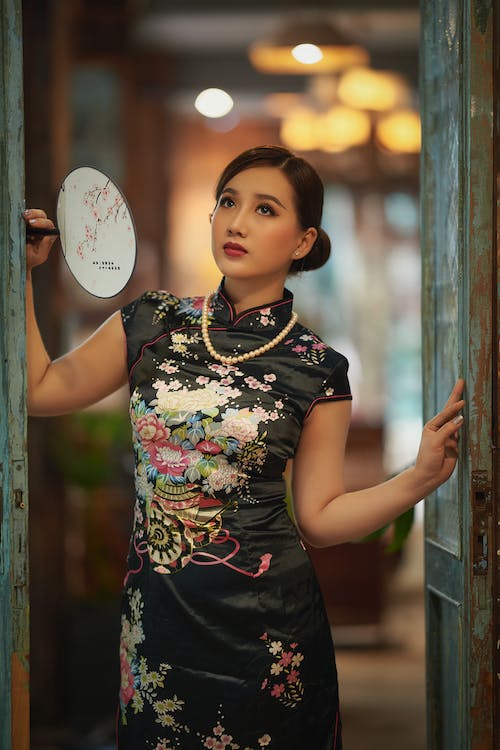 Woman in Black Qipao Dress Standing Beside a Wooden Door