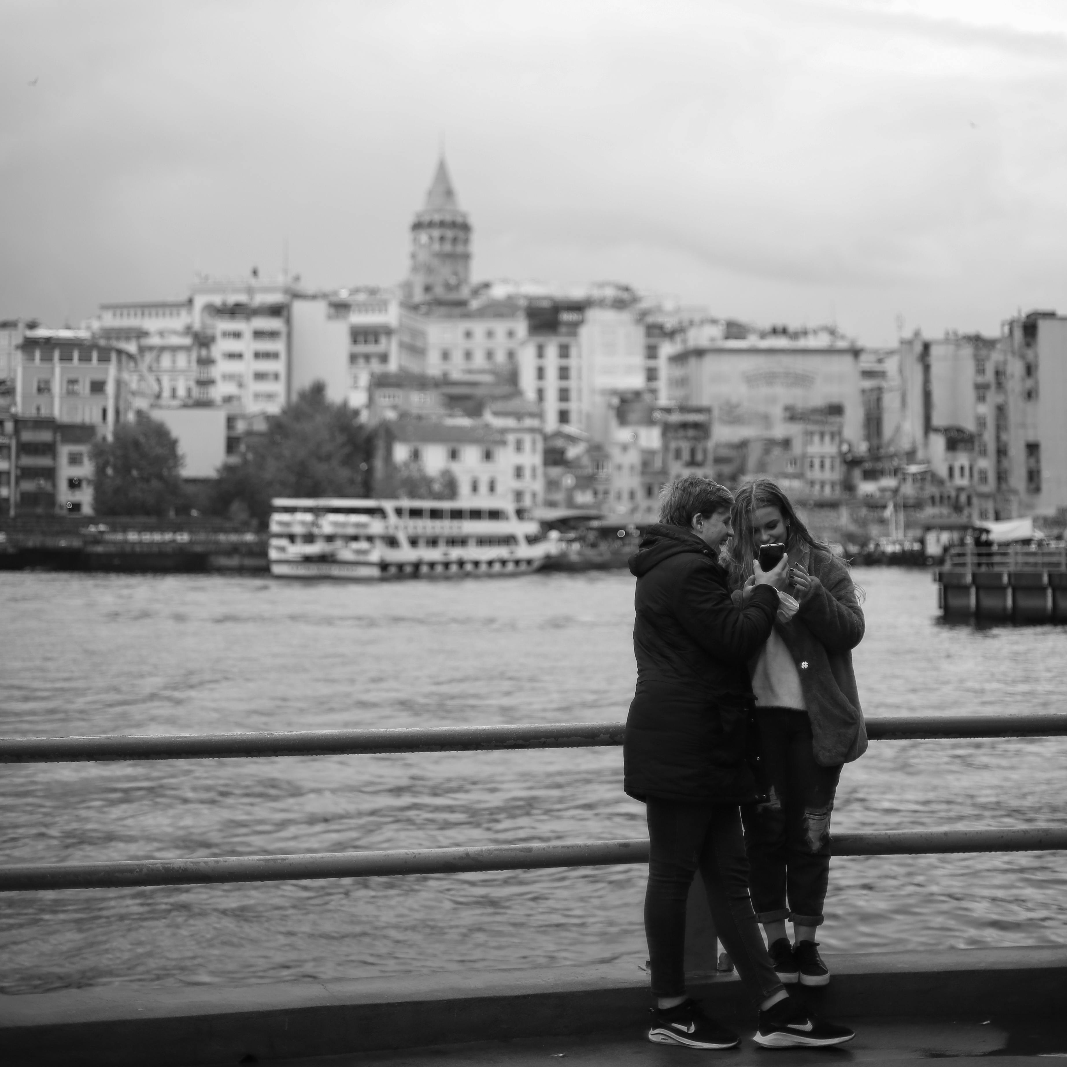 Black and white photo of a couple by the water, with cityscape and Galata Tower in the background.